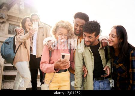 Eine Gruppe fröhlicher Freunde, die sich im Freien ein Smartphone ansehen. Menschen, die digitale Geräte nutzen und in sozialen Medien teilen, treffen zusammen. Stockfoto
