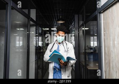 Schwarzer Medizinstudent liest Bücher und bereitet sich auf eine Prüfung vor. Afrikaner in einem medizinischen Gewand und Maske schaut auf die Notizen Stockfoto