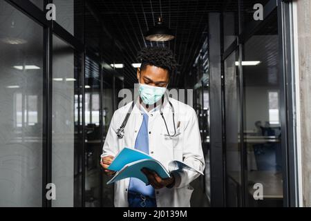 Schwarzer Medizinstudent liest Bücher und bereitet sich auf eine Prüfung vor. Afrikaner in einem medizinischen Gewand und Maske schaut auf die Notizen Stockfoto