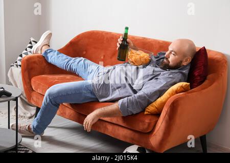 Lustiger fauler Mann, der Bier trinkt und Chips isst, während er zu Hause fernsieht Stockfoto