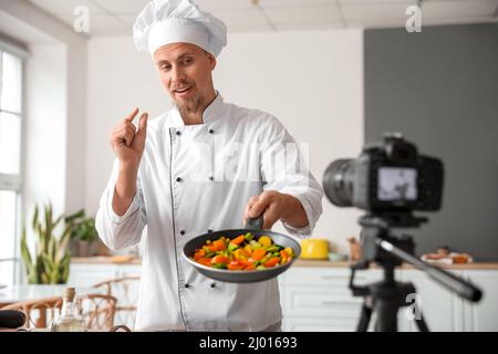 Männlicher Koch mit gebratenem Gemüse, der Video in der Küche aufzeichnet Stockfoto