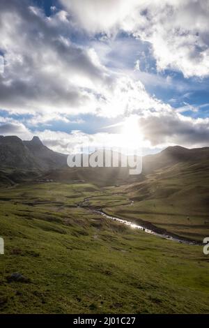 Friedliche Berglandschaft mit einem Fluss durch das Tal mit Tieren grasen in den grünen Wiesen, felsigen Bergen im Hintergrund und die Stockfoto