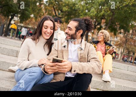 Junge Frau und Mann beobachten etwas Lustiges in der mobilen Zelle. Glückliche Menschen, die telefonieren und Spaß haben Stockfoto
