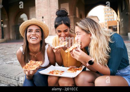 Lustige gemischte Renngruppe von drei Frauen essen Street Food im Freien in der Stadt Straße. Glückliche Freundinnen genießen die Sommerferien zusammen Stockfoto