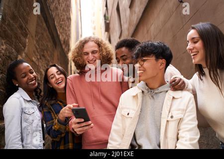 Gruppe von glücklichen jungen Teenagern, die im Freien Smartphone benutzen, während sie sich etwas im Handy ansehen und lachen. Konzept der Technologie, Freundschaft und Stockfoto