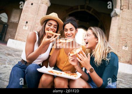 Drei lustige multirassische Frauen essen Pizza im Freien auf der Straße. Glückliche Freundinnen genießen die Sommerferien zusammen europäische Stadt. Jung Stockfoto