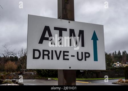 Leicht abgewinkelte Ansicht eines weißen ATM-Drive-Up-Schilds mit einem Richtungspfeil, der auf den Beginn des Drive-Thru zeigt und gegen einen bewölkten Himmel geschossen ist Stockfoto