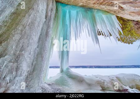 Im Winter hängen große, scharfe blaue Eiszapfen an Felsen neben dem gefrorenen See Stockfoto
