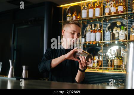 Hübscher junger Barkeeper, der Zutaten eines alkoholischen Cocktails mischt, indem er den Shaker hinter der Theke schüttelt. Stockfoto