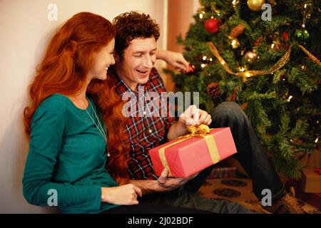 Weihnachten mit einem besonderen Menschen teilen. Aufnahme eines jungen Mannes, der sein weihnachtsgeschenk öffnete, während er mit seiner Freundin saß. Stockfoto