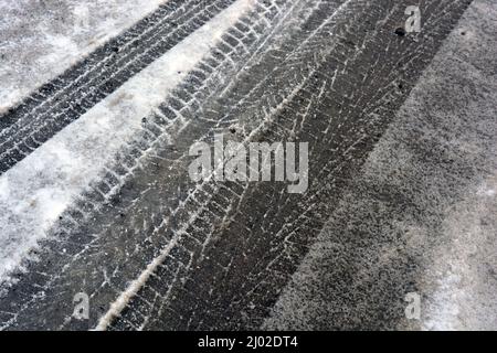 Spuren von Autorädern, vorbei an Autos auf einer verschneiten und schmelzenden Winterstraße, ein interessanter Hintergrund. Stockfoto