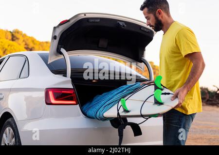 Attraktiver junger Mann mit Bart, der sein Surfbrett aus dem Kofferraum des Autos nimmt, Freizeit- und Hobbys-Konzept Stockfoto