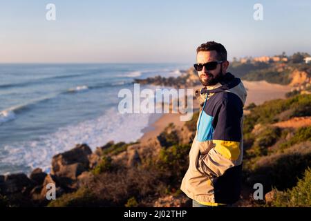 Junger Mann mit Jacke und Sonnenbrille mit Blick auf das Meer von einer Klippe neben dem Strand, Freizeit- und Relax-Konzept, Copy Space for Text Stockfoto