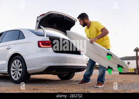 Hübscher junger Mann mit Bart, der sein Surfbrett aus dem Kofferraum des Autos nimmt, Freizeit- und Hobbys-Konzept Stockfoto