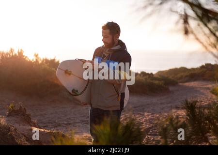 Bärtiger junger Mann in Jacke, der bei Sonnenuntergang durch die Dünen des Strandes mit seinem Surfbrett unter dem Arm, Freizeit- und Hobbys-Konzept, Kopierplatz für läuft Stockfoto