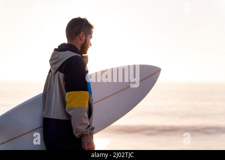 Junger Mann in Jacke mit Blick auf das Meer bei Sonnenuntergang mit seinem Surfbrett unter dem Arm, Freizeit- und Hobbys-Konzept, Copy Space for Text