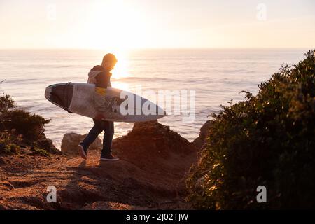 Junger Mann beim Sonnenuntergang mit seinem Surfbrett entlang der Klippen am Meer, Freizeit- und Hobbys-Konzept, Copy Space for Text Stockfoto