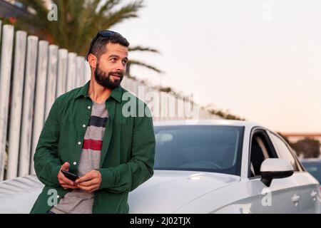 Attraktiver junger bärtiger Mann auf der Motorhaube seines Autos mit Mobiltelefon in der Hand, Freizeit- und Kommunikationstechnikkonzept, Kopierraum für te Stockfoto