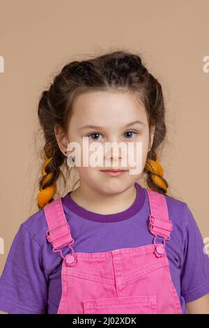 Porträt eines hübschen kleinen Mädchens mit einem leichten Lächeln mit gelben Kanekalon-Zöpfen, mit Augen, die in pinkem Jumpsuit und violettem T-Shirt auf die Kamera schauen Stockfoto