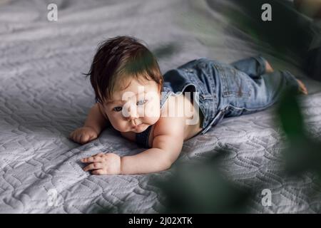 Porträt eines hübschen Babys in Denim-Strampler, der zu Hause auf der Bettdecke ruht, verschwommene grüne Pflanze im Vordergrund. Kleinkind lag auf dem Bauch, erkunden Sie die Welt Stockfoto