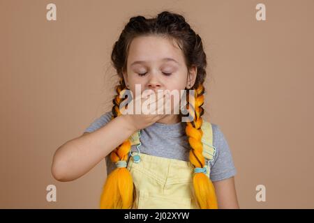 Erschöpftes gähnendes kleines Mädchen mit gelben Kanekalon-Zöpfen, mit geschlossenen Augen, die den Mund schließen, mit Hand im gelben Jumpsuit und grauem T-Shirt auf Beige Stockfoto