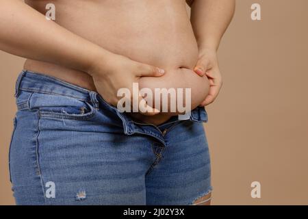 Frau mit großem Bauch und Händen, die Fett in blauen Jeans auf beigem Hintergrund zeigen. Plötzliche Gewichtszunahme. Viszerales Fett. Körper positiv. Wenig eng Stockfoto