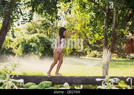 Engagiertes kleines Mädchen, das tagsüber auf dem Holzweg balanciert und mit Wassersprinklern, Haus und grünen Bäumen im Hintergrund wegschaut. Träumen Stockfoto