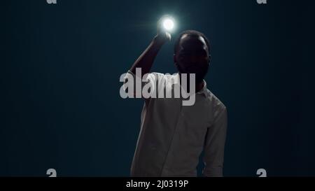 Junge Person, die im Dunkeln sitzt und mit Taschenlampe nach Beweisen sucht. Männlicher Detektiv riskiert und hält Laterne, um gruselige Geräusche zu untersuchen und zu untersuchen. Stockfoto