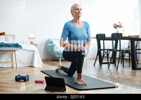 Ältere Frau, die mit einem Online-Trainer in ihrem Wohnzimmer Übungen macht. Stockfoto