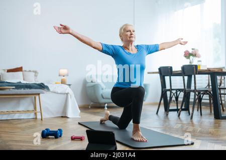 Ältere Frau, die mit einem Online-Trainer in ihrem Wohnzimmer Übungen macht. Stockfoto