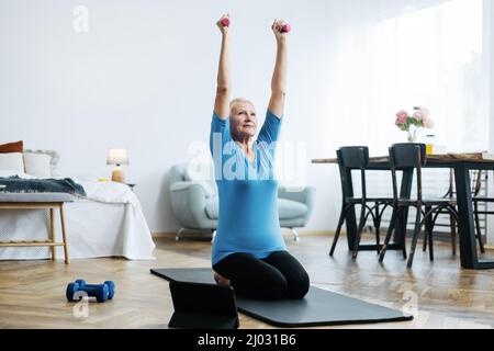 Ältere Frau macht Übungen mit Kurzhanteln in der Nähe des Laptops im Wohnzimmer. Stockfoto