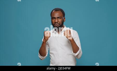 Frustrierte Person, die Fäuste zusammenhält und Kampfdrohungen vor der Kamera macht. Aggressiver Mann mit Stärke, der im Studio Boxen vorführt und Schläge vorbereitet, um negative Wut zu werben. Stockfoto