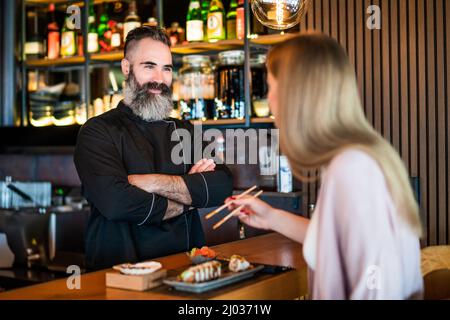 Im Sushi-Restaurant isst und verkostet eine junge Frau Sushi mit Essstäbchen. Sushi-Meisterin wartet auf ihre Meinung. Stockfoto