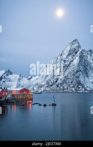 Rote Rorbu-Hütten und majestätischer Olstind-Gipfel, der während der Winterdämmerung vom Mond beleuchtet wird, Hamnoy, Nordland County, Lofoten Islands, Norwegen, Skandinavien, Europa Stockfoto