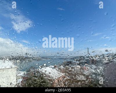 Regentropfen, Regentropfen auf der Fensterscheibe über verschwommenem Himmel und Blick auf die Stadt unter Schnee. Wassertropfen auf dem Fenster vor verschwommenem Hintergrund oder Oberfläche mit Häusern wh Stockfoto