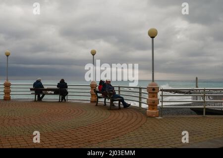 Menschen sitzen auf Bänken am Wasser an einem bewölkten Wintertag mit rauem Meer, Bordighera, Imperia, Ligurien, Italien Stockfoto