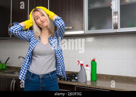 Die junge blonde Frau in der Küche hält ihren Kopf in gelben Handschuhen und zeigt emotional Ärger, Unzufriedenheit mit der anstehenden Reinigung Stockfoto