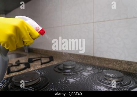 Nahaufnahme der Hand einer Frau in gelben Handschuhen, die Reinigungsmittel auf den eingebauten Küchenherd stoßen Stockfoto