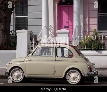 Beigefarbener Oldtimer Fiat 500, der vor einem Haus mit einer rosafarbenen Tür in einer Wohnstraße in Notting Hill, West-London, Großbritannien, geparkt ist. Stockfoto