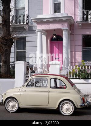 Beigefarbener Oldtimer Fiat 500, der vor einem Haus mit einer rosafarbenen Tür in einer Wohnstraße in Notting Hill, West-London, Großbritannien, geparkt ist. Stockfoto