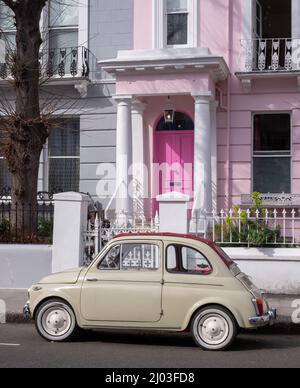 Beigefarbener Oldtimer Fiat 500, der vor einem Haus mit einer rosafarbenen Tür in einer Wohnstraße in Notting Hill, West-London, Großbritannien, geparkt ist. Stockfoto