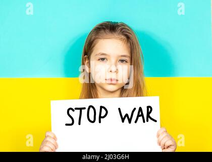 Ein Mädchen weint und hält ein STOP-WAR-Plakat in den Händen vor dem Hintergrund der ukrainischen Flagge. Beendet den Krieg in der Ukraine. Das Konzept des Friedens Stockfoto