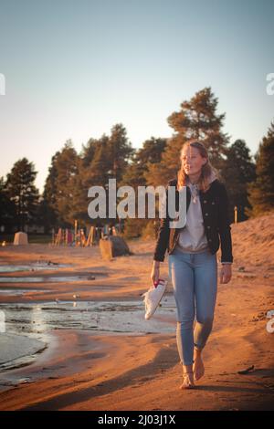 Lächelnder, junger, brünetiger Athlet geht barfuß an einem Sandstrand in Jeans und schwarzer Bluse und genießt den Sonnenuntergang und die letzten warmen Strahlen. Sotkamo, Finla Stockfoto