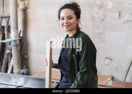 Junger Zimmermann mit Holzbrett und lächelnd in der Werkstatt vor der Kamera Stockfoto