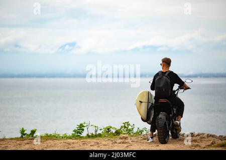 Junger Mann sitzt auf einem Motorrad mit einem Surfbrett. Hochwertige Foto Stockfoto