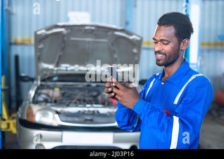 Happy Car Mechanic beschäftigt mit Handy in der Garage vor dem Reparaturauto mit geöffneter Haube - Konzept der Verwendung von Technologie, Kunden online zu finden Stockfoto