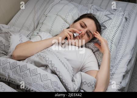 Eine Frau mit Rhinitis quietscht eine Kochsalzlösung oder tropft Vasokonstriktor in die im Bett liegende Nase, Draufsicht. Stockfoto