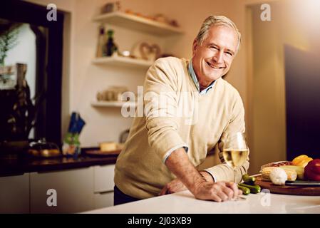 Wein ist mein Lieblingsgericht. Aufnahme eines älteren Mannes, der ein Glas Wein genießt, während er in seiner Küche das Abendessen zubereitet. Stockfoto