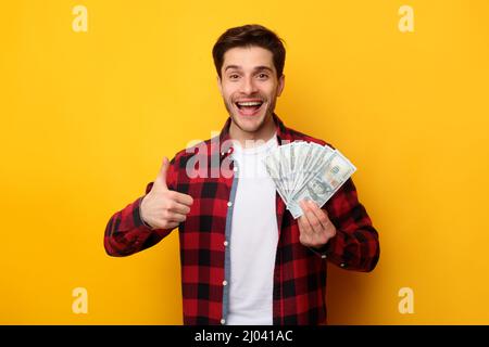 Aufgeregter Mann, der Geld in bar hält und Daumen nach oben Stockfoto