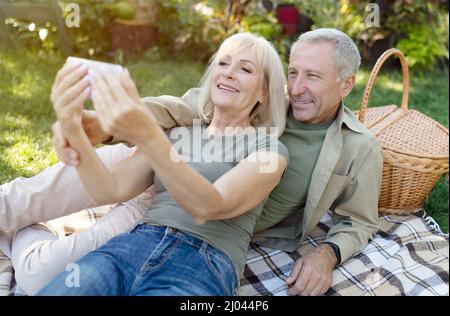 Glückliche ältere Ehepartner telefonieren mit dem Smartphone, während sie sich am warmen Frühlingstag im Garten ausruhen und picknicken Stockfoto